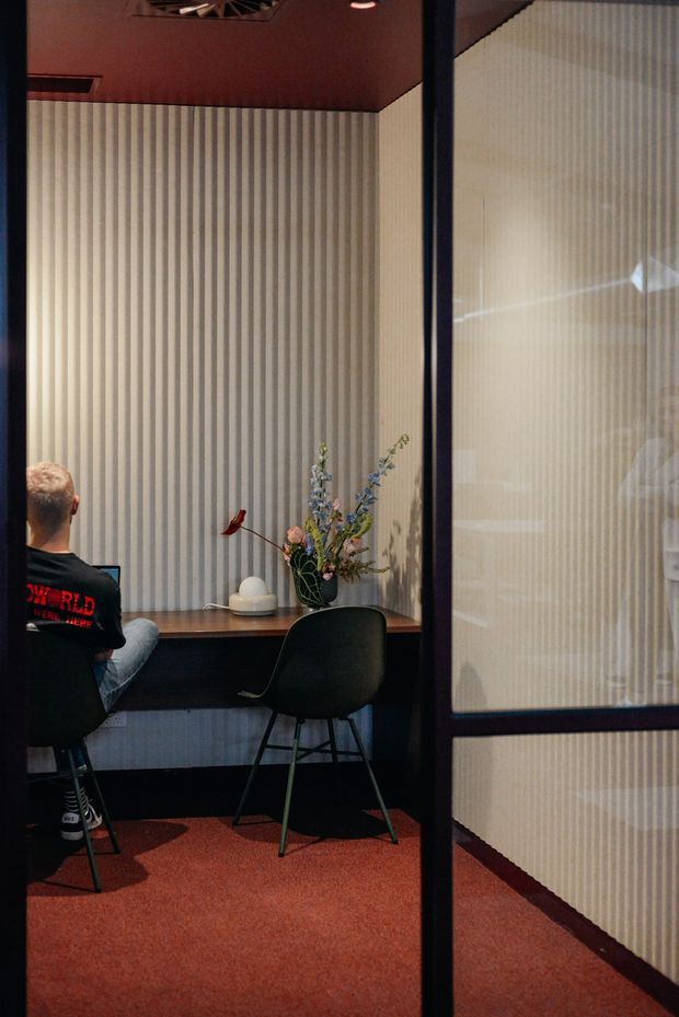 A compact, enclosed workspace or meeting booth viewed through a dark-framed glazed partition. The room has vertically ribbed wall panels, a warm terracotta-red ceiling and carpet, a built-in dark timber desk, black chairs, decorative objects and a small floral arrangement. A person is seated at the desk, and the glazing reflects or reveals the adjacent space.