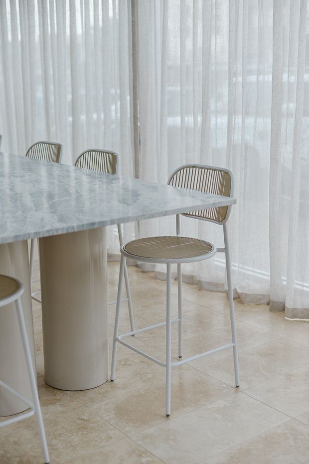 Bright, refined dining area featuring a pale marble or stone tabletop on a cylindrical pedestal base, surrounded by slim white metal bar-height chairs with curved slatted backs and woven-looking seats. Full-height sheer white curtains diffuse natural light, while the floor is finished in large warm beige stone-look tiles.