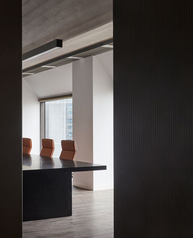 A refined contemporary dining or meeting area viewed through a dark vertical slatted partition. A long black built-in table with warm brown upholstered chairs sits beside a large window, while white walls, pale timber flooring, exposed ceiling panels, and integrated linear lighting create a restrained architectural interior.