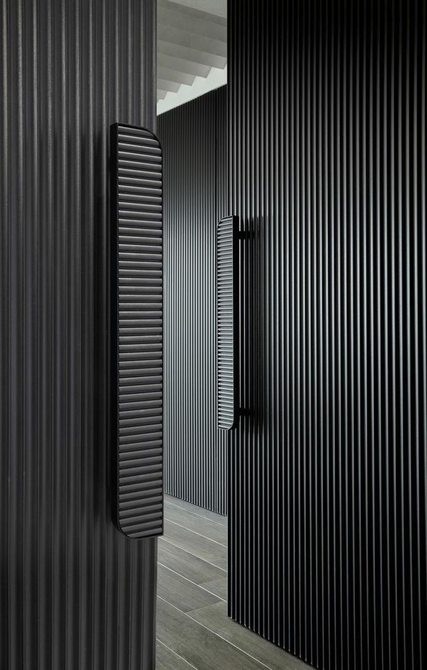 A narrow, dramatic interior passage lined with vertical black ribbed wall panels and matching elongated ribbed door handles. Grey wood-look flooring runs through the corridor, while a white ceiling with exposed or stepped beams is visible at the upper opening. The image is highly monochromatic, emphasizing strong vertical rhythm, texture, and refined modern detailing.