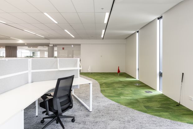Bright contemporary open-plan office interior with white workstations and partition screens in the foreground, a black ergonomic office chair, and a broad green patterned carpet zone extending beside full-height windows. The space has a suspended white acoustic tile ceiling with linear recessed lighting, minimal white walls, and a clean professional fit-out.
