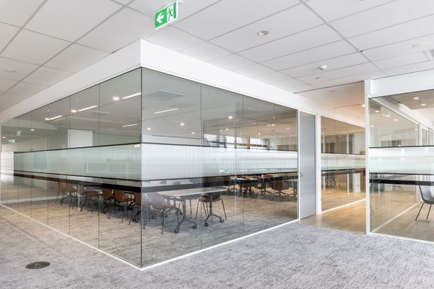 Bright contemporary commercial office interior with glazed meeting or collaboration rooms enclosed by full-height glass partitions. The space features a suspended white acoustic tile ceiling, recessed lighting, pale grey carpet, white framing, and rows of movable tables and chairs visible through the glazing. Frosted horizontal bands provide privacy, and an illuminated emergency exit sign is visible overhead.
