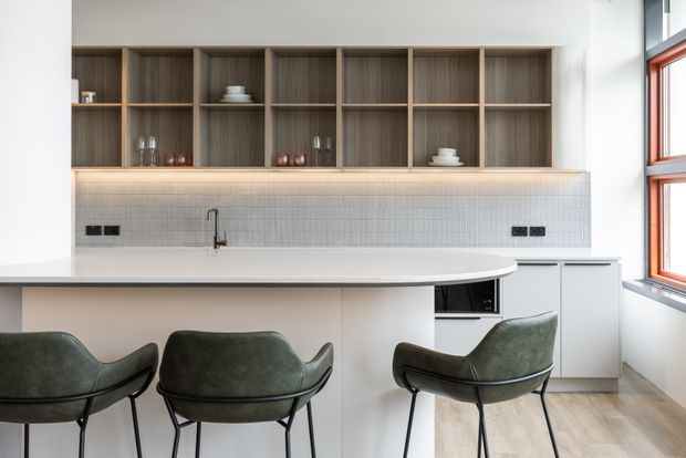 A bright contemporary kitchen with a long white island and three dark green upholstered bar stools. The rear wall features open dark timber shelving, pale built-in cabinetry, a fine light mosaic-tile backsplash, under-shelf lighting, and a stainless tap and sink. Warm timber flooring and a large red-framed window add contrast.