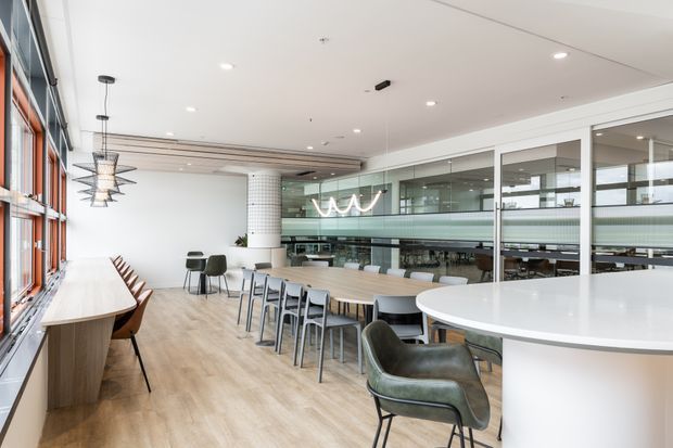 Bright contemporary commercial dining and breakout area with a long communal timber table, integrated window-side bench seating, modern pendant lights, a curved white counter, and glazed meeting or office spaces beyond. The space has pale timber-look flooring, white walls and ceiling, expansive glazing, and a clean open-plan workplace or hospitality fit-out.