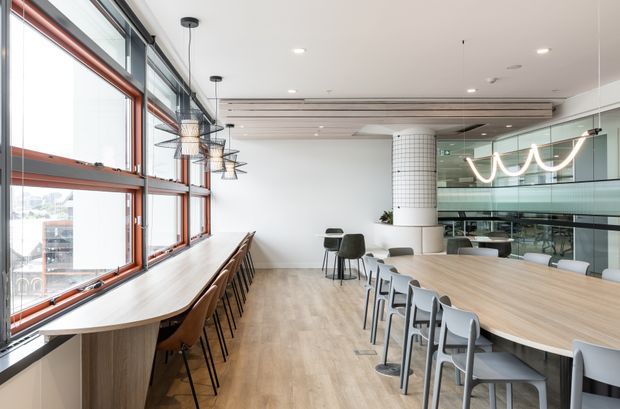 Bright contemporary communal dining and collaboration space with long timber tables, rows of chairs, a window-side bench, exposed red-toned window frames, pendant lighting, a tiled cylindrical column, and glazed views into an adjacent office area.