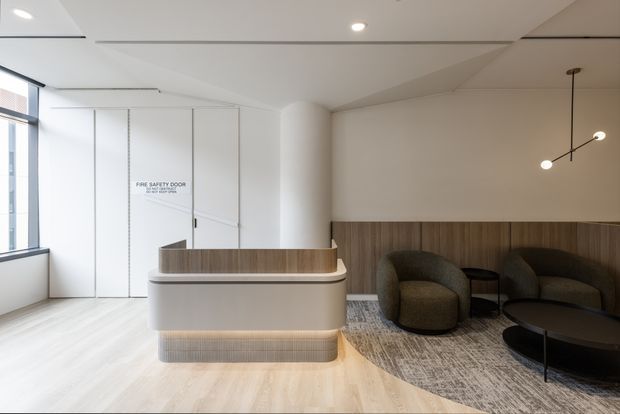 A refined contemporary reception lobby with a curved built-in reception desk, integrated warm lighting, wood-panelled feature wall, sculptural pendant light, and a small lounge seating area. The space is predominantly white and light timber with dark upholstered furniture and a textured area rug.