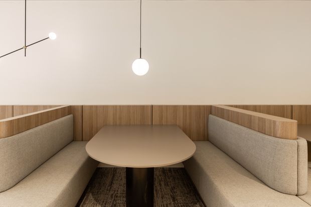 A refined built-in booth seating area with a rounded rectangular dining table, upholstered beige banquettes, warm timber privacy screens, and minimalist pendant lighting. The space has a calm, contemporary hospitality or dining character with neutral finishes and a softly lit atmosphere.