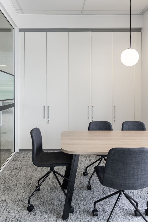 A bright, minimalist office meeting room with a long light timber meeting table, dark upholstered swivel chairs, full-height white storage cabinetry, grey carpet, glass partitions, and a simple spherical pendant light. The space has a clean contemporary commercial fit-out with strong vertical cabinetry lines and abundant daylight.
