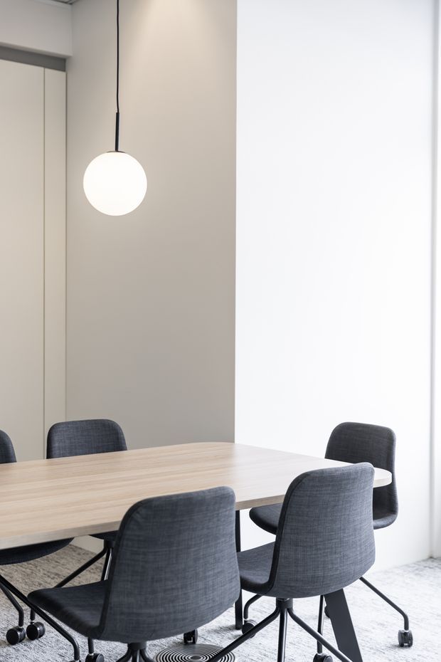 Bright minimalist dining or meeting area with a long light timber table, dark upholstered wheeled chairs, pale walls, a large white sheer window covering, and a simple spherical pendant light. A mirrored or reflective panel is visible along the left wall, extending the view of the room.