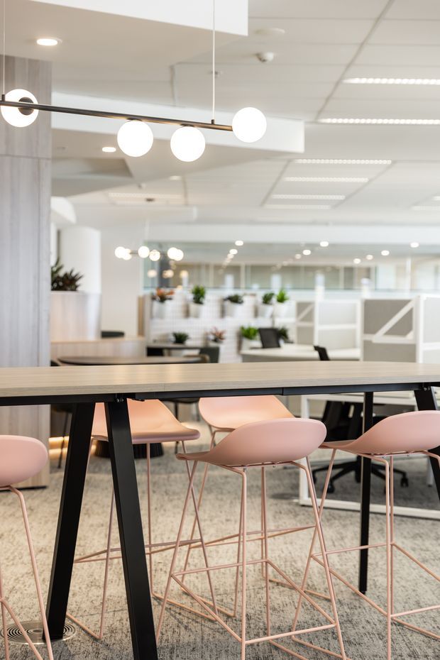 A bright contemporary open-plan office interior with a long light-wood communal table on black angled legs, surrounded by pale pink bar stools and chairs. Suspended globe pendant lighting, white acoustic ceiling tiles, carpeted flooring, planted work areas, and glass-partitioned workspaces create a modern collaborative workplace setting.