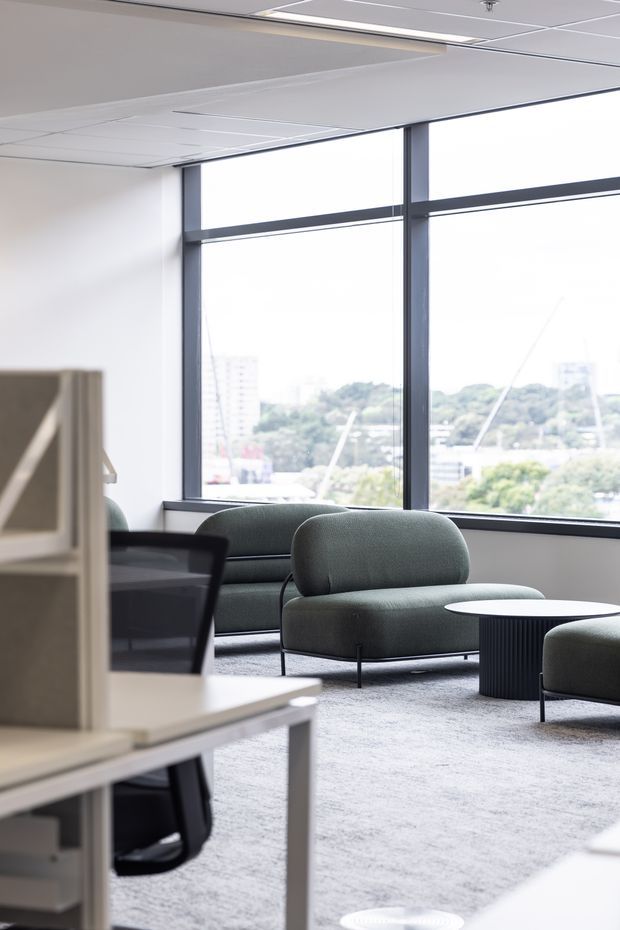 A bright contemporary commercial lounge or breakout area beside large floor-to-ceiling windows. The space contains rounded upholstered green-grey lounge chairs and ottomans, a dark ribbed round coffee table, pale carpet, white walls, and a suspended acoustic ceiling with integrated linear lighting. Workstations and office furniture are visible in the foreground, while an urban landscape appears through the glazing.