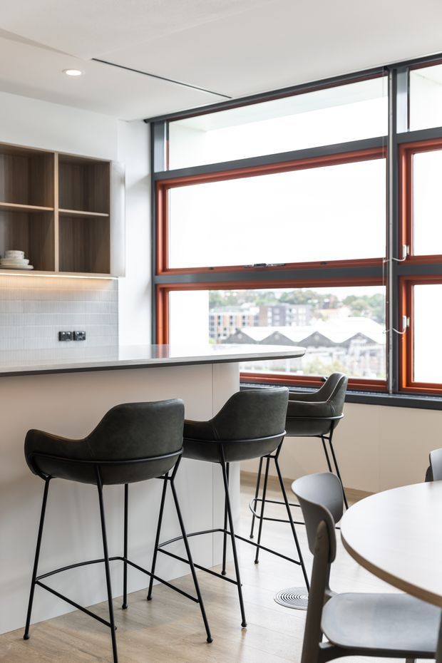 Bright contemporary kitchen and dining area with a peninsula breakfast bar, dark upholstered bar stools, built-in timber shelving, subtly textured tiled splashback, and large industrial-style framed windows with red-brown timber reveals. A round dining table is partially visible in the foreground.