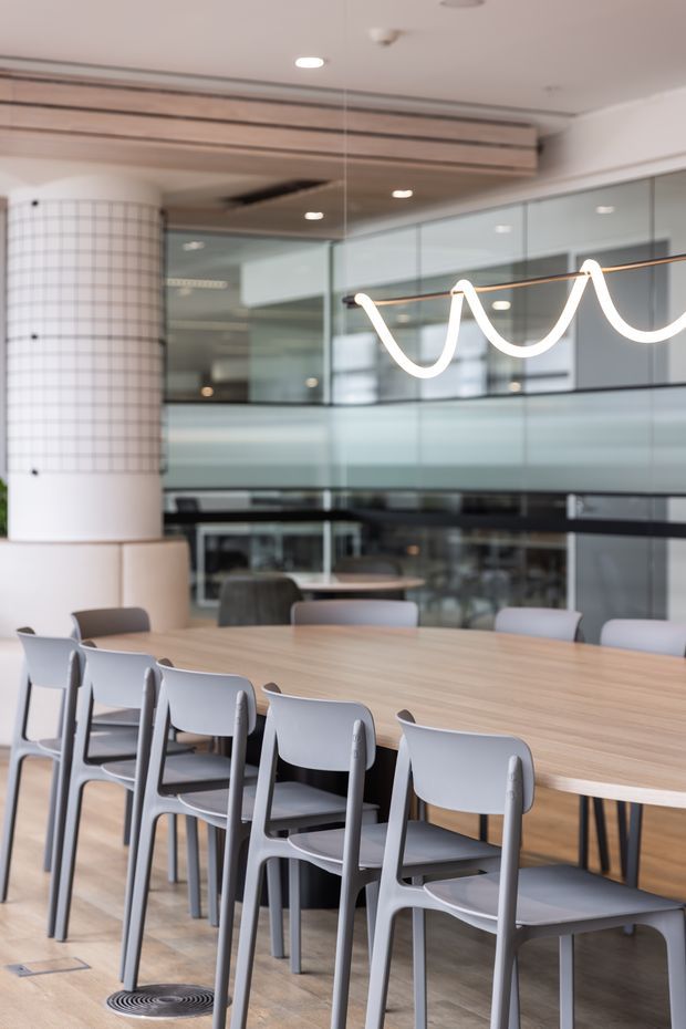 Contemporary communal dining area with a long light-wood table and rows of grey chairs. The space features a tiled cylindrical column, extensive interior glazing, reflective glass partitions, a timber-lined ceiling detail, and a distinctive linear pendant light with looping white forms.