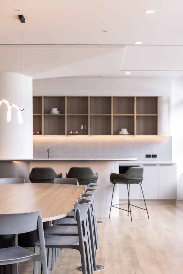 Bright, refined open-plan kitchen and dining area with a large light timber dining table, grey dining chairs, a kitchen island with bar stools, white cabinetry, dark timber open shelving, under-shelf lighting, and a pale tiled splashback. Recessed ceiling lighting and a sculptural pendant add contemporary detail.