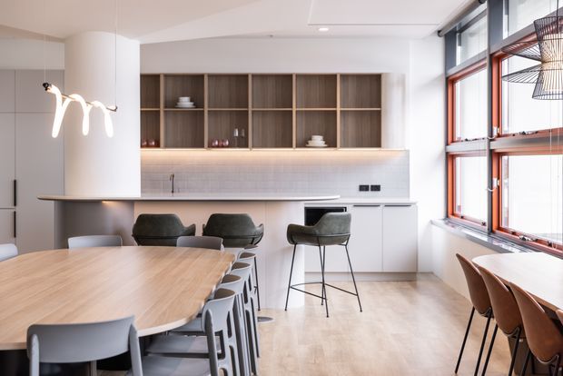 Bright open-plan kitchen and dining area with a large light timber dining table, contemporary seating, integrated white cabinetry, a timber open display-shelving wall, pale tiled splashback, and a slim kitchen island with stools. Full-height windows with reddish timber frames line the right side, while sculptural pendant lighting and warm under-cabinet illumination add visual interest.