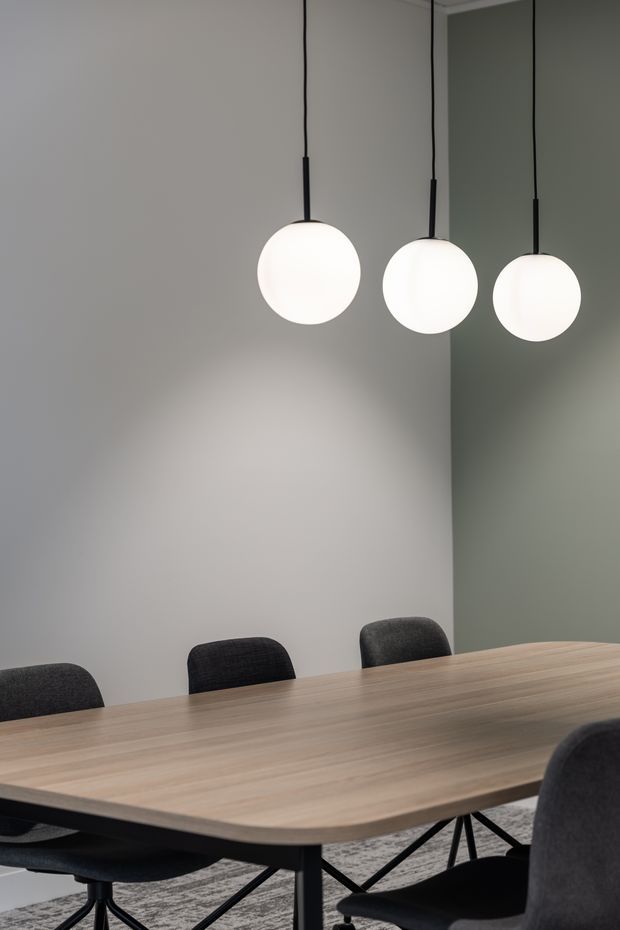 A minimalist dining room with a large light timber dining table, dark upholstered dining chairs, and three spherical pendant lights suspended over the table. The room has plain light grey and muted green walls, a neutral patterned rug, and a calm contemporary aesthetic.