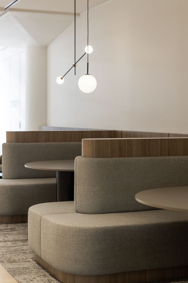 A refined contemporary dining or hospitality interior with curved upholstered booth seating, round tables, and warm wood privacy screens. Minimal white walls and ceiling are complemented by sculptural globe pendant lights, creating a calm, understated, softly lit setting.