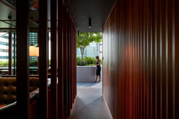 A narrow, sophisticated interior passage framed by rich dark timber vertical slats. The corridor has a smooth dark floor, a simple charcoal ceiling with recessed linear lighting, and glimpses of a refined lounge or reception area through the slatted partition on the left. It opens at the far end to a landscaped courtyard with a raised planter and urban buildings beyond. The atmosphere is moody, refined, and highly architectural.