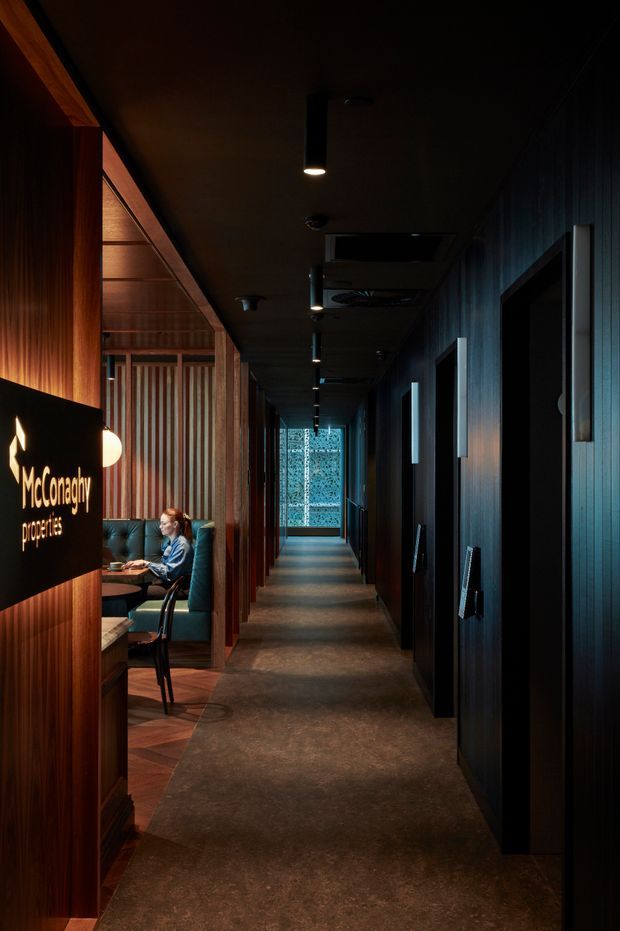 A dimly lit commercial hospitality corridor with dark timber wall panelling, black doors, warm concealed and downlighting, and a textured dark floor. On the left, a partially open booth seating area with a diner and a branded McConaghy Properties sign is visible. The corridor leads toward a patterned glazed or illuminated feature at the far end, creating a dramatic, refined atmosphere.