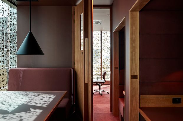 A compact, richly detailed interior corridor connecting a dining nook and a home office. Dark timber doors, wall panelling and built-in joinery contrast with muted mauve walls and red carpeting. A black cone pendant hangs above a built-in upholstered bench and dining table, while patterned glazing casts dappled light across the surfaces. An office chair and desk are visible through the doorway in the background.