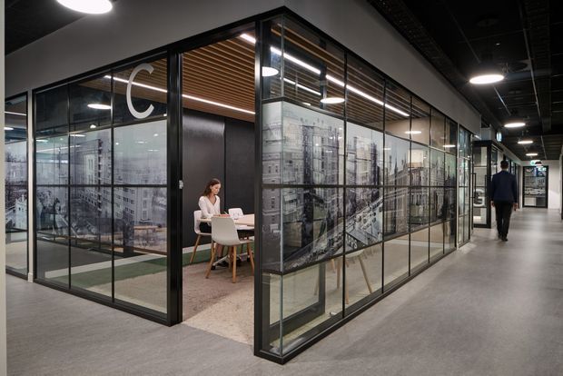 A contemporary commercial office interior viewed from a circulation corridor. A glazed meeting room or private workspace occupies the centre, enclosed by black-framed glass partitions with large monochrome architectural cityscape graphics. Inside are a small round table, chairs, a seated person using a laptop, carpet flooring, and a timber slatted ceiling feature. The surrounding office has dark exposed ceiling services, recessed and circular ceiling lights, and polished grey flooring.