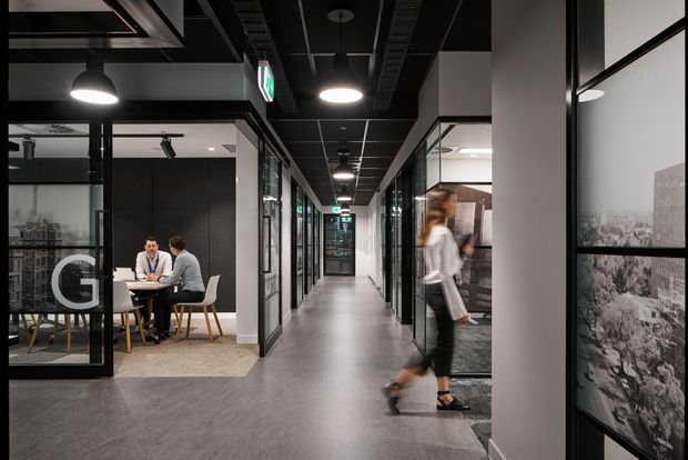 A contemporary commercial office interior viewed along a long central corridor. Glazed meeting rooms line both sides, with people seated around a small table in one room and a blurred person walking through the corridor. The space has a dark exposed-services ceiling grid, suspended circular pendant lights, black-framed glass partitions, grey flooring, white structural columns, and monochrome photographic panels along the right wall.