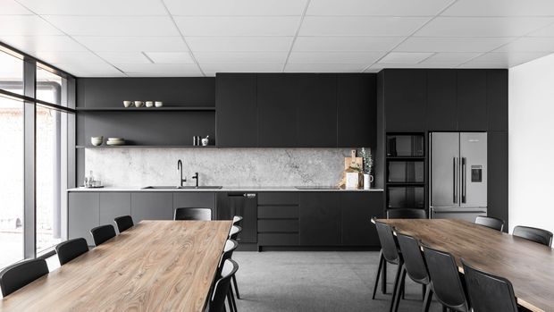 A contemporary commercial-style kitchen and dining area with extensive matte-black cabinetry, open shelving, a pale stone-look splashback, integrated appliances, and two large timber dining tables with black chairs. The space has full-height glazing on the left, a suspended white grid ceiling, and a polished industrial-modern appearance.