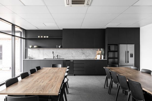 Contemporary open-plan commercial kitchen and dining area with matte black full-height cabinetry, open display shelving, a pale marble-look splashback, stainless-steel appliances, and two large timber dining tables surrounded by black chairs. Full-height glazing lines the left side, while a suspended acoustic tile ceiling and exposed air-conditioning unit contribute to the workplace or hospitality setting.