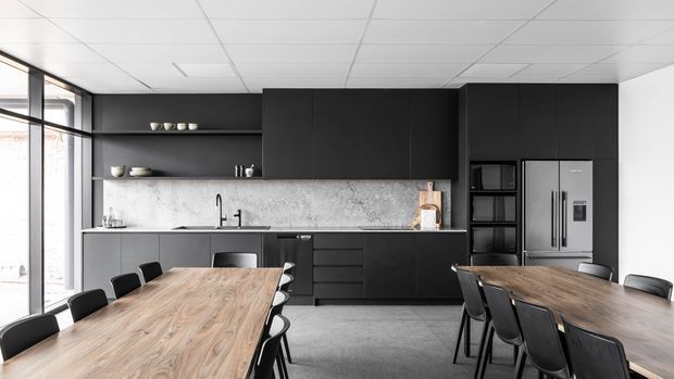 A contemporary open-plan kitchen and dining area with extensive matte black cabinetry, a pale stone-look splashback, integrated appliances, and two large timber dining tables with black chairs. Full-height glazing lines the left side, and the space has a clean suspended grid ceiling and polished concrete-look floor.