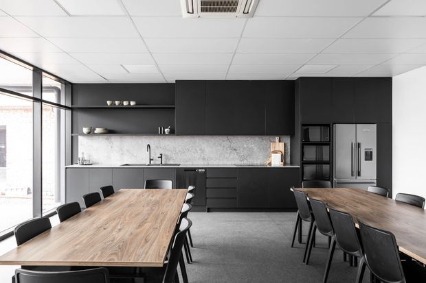 A contemporary open-plan commercial-style kitchen and dining area with extensive matte-black cabinetry, open shelving, a pale stone-patterned splashback, integrated appliances, and a long timber dining table surrounded by black chairs. Floor-to-ceiling glazing lines the left side, while a suspended acoustic tile ceiling and exposed air-conditioning unit complete the functional fit-out.