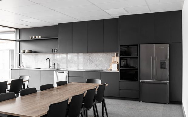 A contemporary open-plan kitchen and dining area with extensive matte dark cabinetry, integrated black appliances, a large stainless-steel refrigerator, pale stone-look splashback, light grey cabinetry, and a long timber dining table with black chairs. A large window provides natural light, while open shelving displays ceramics. The space has a clean, refined commercial or workplace-style fit-out.