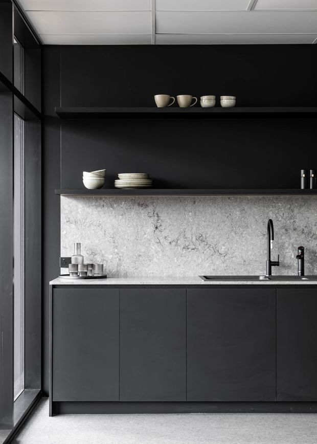 A compact, highly refined dark kitchen with matte black full-height cabinetry, open display shelving, a pale veined stone splashback and benchtop, and black minimalist plumbing fixtures. Large dark-framed glazing runs along the left side, while a suspended panel ceiling and pale polished floor provide contrast. Ceramic vessels and tableware add subtle warmth to the monochrome scheme.
