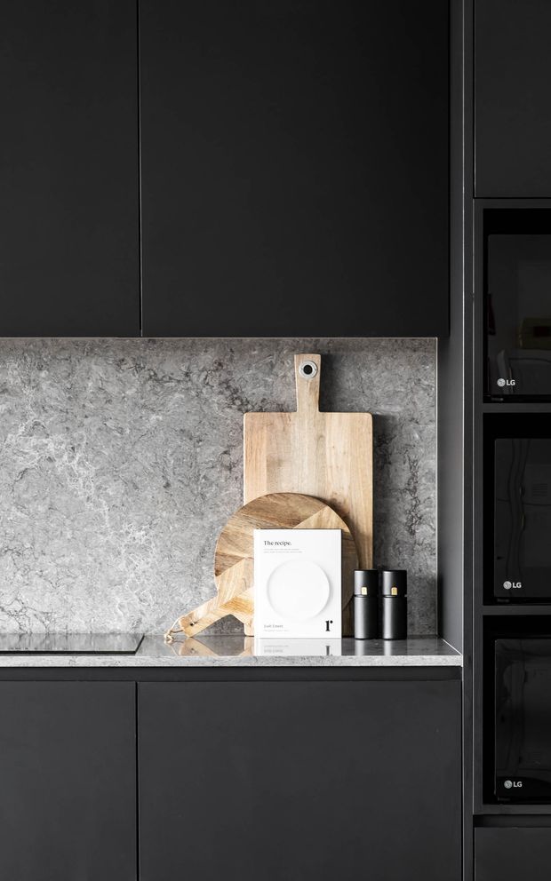Close-up view of a sophisticated dark kitchen with matte charcoal-black cabinetry, a grey veined stone backsplash and matching benchtop, and integrated black appliances. Wooden chopping boards, black grinders, and a small cookbook provide restrained styling accents on the counter.