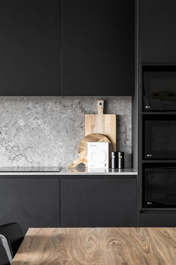 Close-up of a dark, contemporary kitchen with matte black flat-panel cabinetry, a grey veined stone splashback and benchtop, integrated black appliances, and a light-to-mid timber dining table in the foreground. Wooden cutting boards and minimal accessories sit against the splashback.