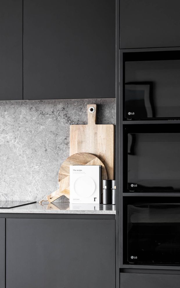 Close-up of a sleek, dark-toned kitchen with matte charcoal cabinetry, a grey veined stone backsplash and benchtop, and a tall bank of integrated black LG appliances. Wooden chopping boards and a cookbook provide subtle styling accents on the counter.