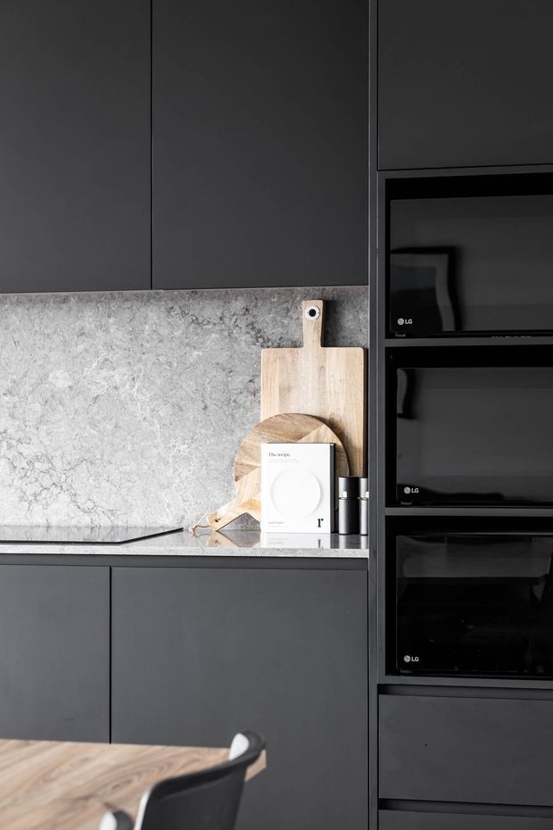 A close-up view of a dark, minimalist kitchen featuring matte charcoal cabinetry, a pale grey veined stone splashback and benchtop, and a tall bank of integrated black appliances. Wooden cutting boards and a cookbook sit on the benchtop, while part of a timber dining table and black chair is visible in the foreground.