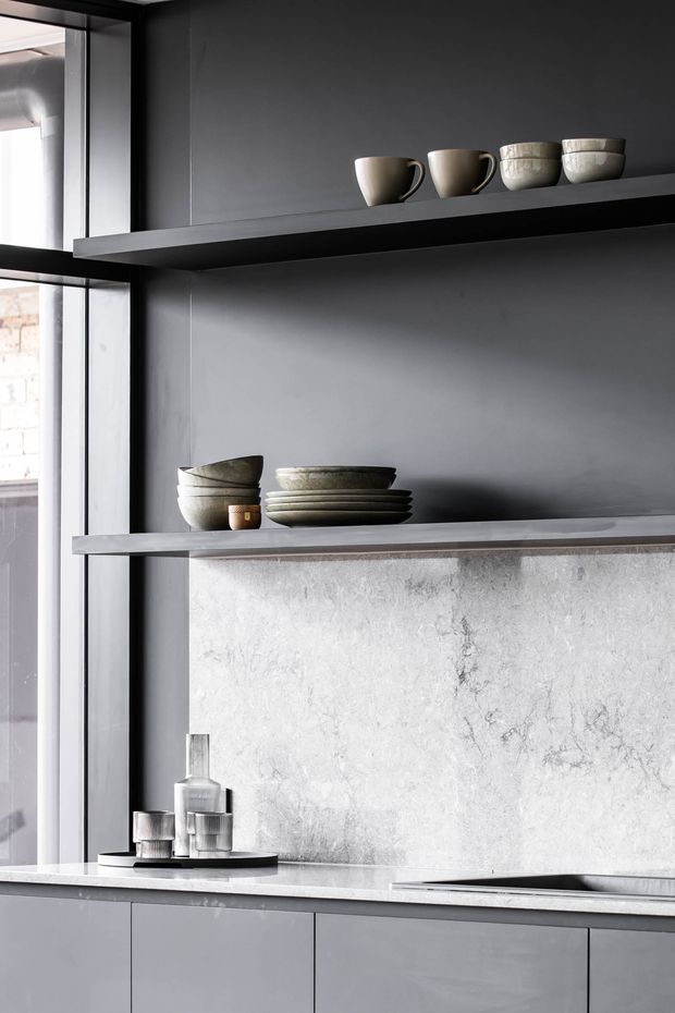 A minimalist contemporary kitchen with dark grey open shelving, grey cabinetry, a pale stone benchtop and matching textured stone splashback. Neutral ceramic cups, bowls and plates are arranged on the shelves, with a stainless steel sink and simple metal accessories below. Tall black-framed glazing brings in bright natural light from the left.