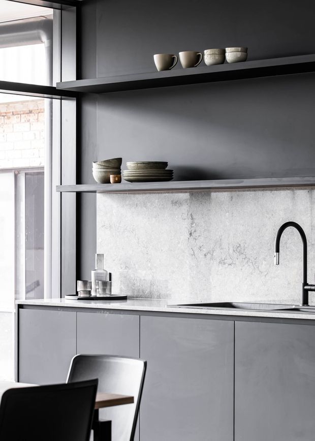 A minimalist modern kitchen in a monochromatic grey palette, featuring flat-panel cabinetry, a pale stone benchtop and matching textured splashback, open dark shelving, a black gooseneck tap, and black-framed industrial-style glazing. Neutral ceramic tableware is displayed on the shelves.