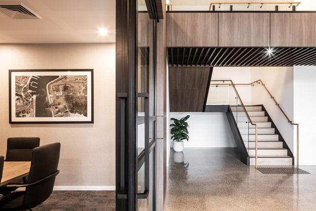 Interior view of a refined contemporary commercial or residential entry and stairway area. A glazed black-framed partition separates a furnished meeting or dining space from a polished concrete-floored stair hall. The stair features white risers, dark side walls, timber handrails, and a dramatic black slatted ceiling beneath a wood-toned upper volume. Neutral walls, framed artwork, recessed lighting, and a potted plant complete the space.