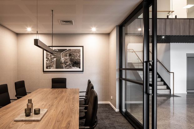 A refined commercial meeting room with a large timber conference table, black upholstered chairs, framed artwork, pendant and recessed lighting, and carpeted flooring. Full-height black-framed glazed partitions reveal an adjacent contemporary stairway and mezzanine area with dark timber or wood-look cladding and polished concrete flooring.