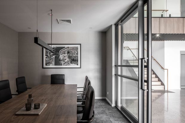 A contemporary office meeting room with a long dark timber conference table, black upholstered chairs, grey carpet, neutral walls, and a linear suspended pendant light. Full-height black-framed glazing looks into a bright stairwell with a mezzanine or upper-level walkway and timber handrail. A framed monochrome artwork hangs on the rear wall.