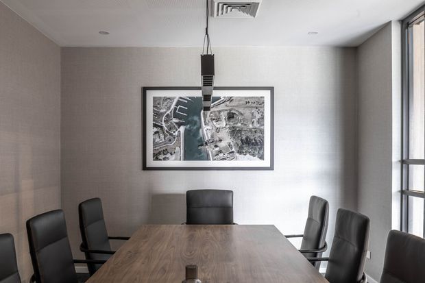 Contemporary meeting or dining room with a large rectangular timber table surrounded by black leather chairs. The room features textured neutral walls, a suspended linear pendant light, a framed aerial artwork, recessed ceiling fixtures, and tall black-framed glazing along one side.