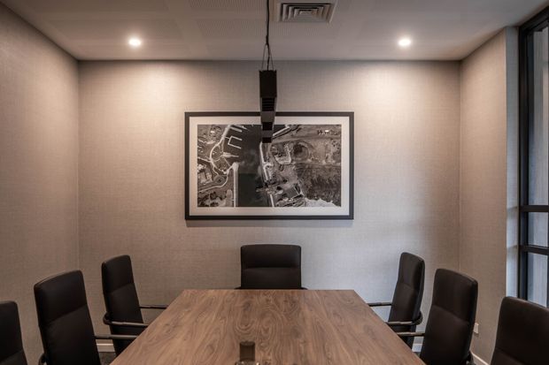 A contemporary boardroom or meeting room with a large rectangular wood meeting table surrounded by black upholstered chairs. The room has warm textured neutral walls, recessed ceiling lighting, visible HVAC, a suspended linear pendant fixture, a framed monochrome artwork, and tall dark-framed glazing along one side.