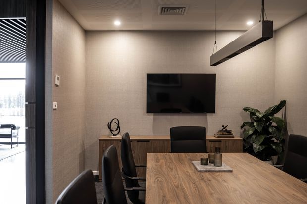 A refined private meeting room or home office with a large timber meeting table, dark upholstered chairs, a wall-mounted television or display, and a low timber sideboard. Textured neutral walls, recessed ceiling lights, and a suspended linear pendant create a warm, professional atmosphere. A leafy indoor plant and a partially visible glazed opening add contrast.