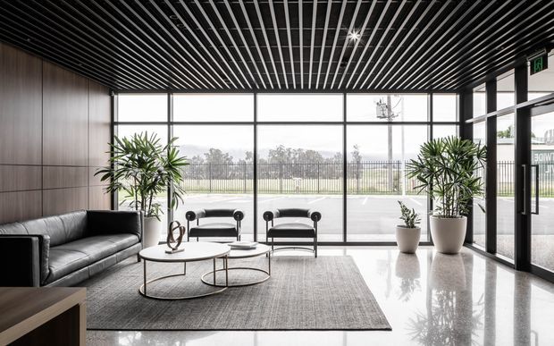 A contemporary commercial lobby or reception area with full-height glazing, dark linear slatted ceiling, timber wall panelling, polished light flooring, black leather seating, round nesting coffee tables, indoor plants, and a large neutral rug. The space overlooks an exterior parking or landscaped area.