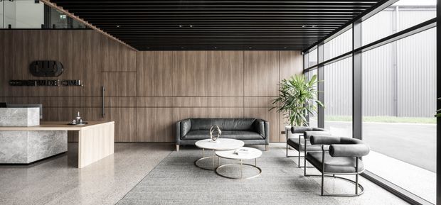 A refined contemporary commercial lobby or reception area with full-height glazing, dark linear ceiling slats, warm timber wall panelling, a stone reception desk, and a lounge seating arrangement with leather sofas and round coffee tables. The space has a clean, spacious, high-end appearance with strong natural light.