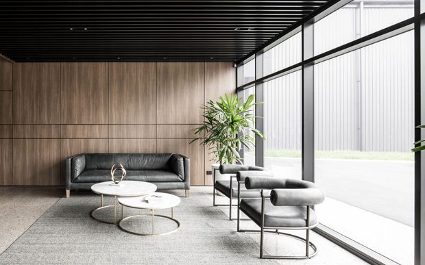 A refined contemporary lounge or reception area with a dark slatted ceiling, full-height black-framed glazing, warm vertical timber wall panelling, and polished concrete flooring. A charcoal sofa and matching lounge chairs surround nested round coffee tables on a large neutral rug, with indoor plants adding greenery. The space has strong natural light and an industrial-modern material palette.