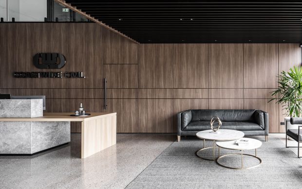 Contemporary commercial reception lobby with a dark timber-look feature wall, black linear ceiling, stone reception desk, polished concrete or terrazzo flooring, and a black leather sofa with round nesting coffee tables. A company logo is mounted on the feature wall, and indoor planting adds a natural accent.