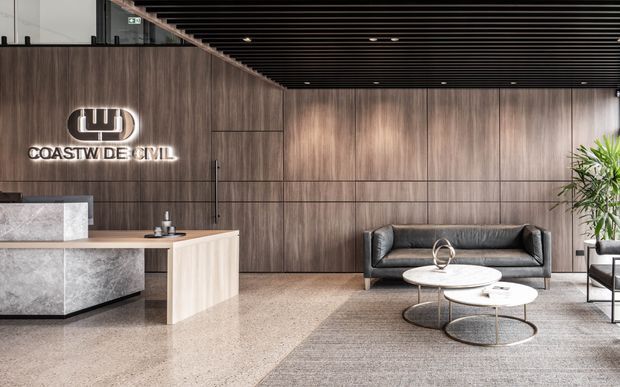 A refined commercial reception lobby with a large stone reception desk, dark timber wall panelling, illuminated Coastwide Civil signage, a black slatted ceiling, and a contemporary lounge seating area with round coffee tables. The space has a neutral, sophisticated palette and polished finishes.
