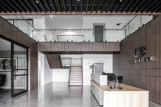 A contemporary commercial reception area with a timber-clad mezzanine and feature wall, polished concrete flooring, black-framed glass partitions, and a light timber reception desk. A stair leads to an upper-level workspace enclosed by glass balustrades. The exposed dark ceiling structure and restrained palette create an industrial, refined office entrance.